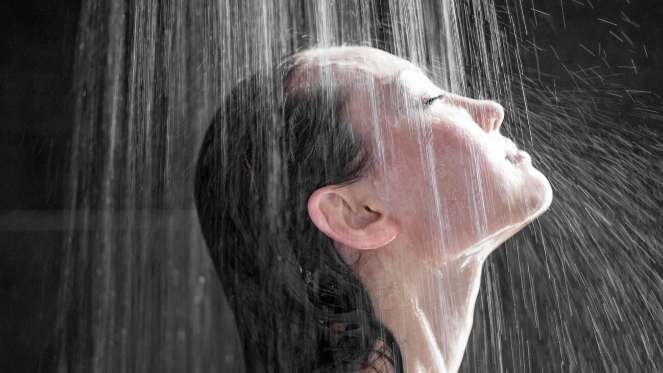Une femme se tient sous la douche et place son visage sous le jet de la douche.
