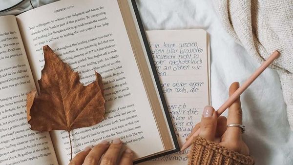Open boeken, daarop een herfstblad en een hand die een potlood vasthoudt.