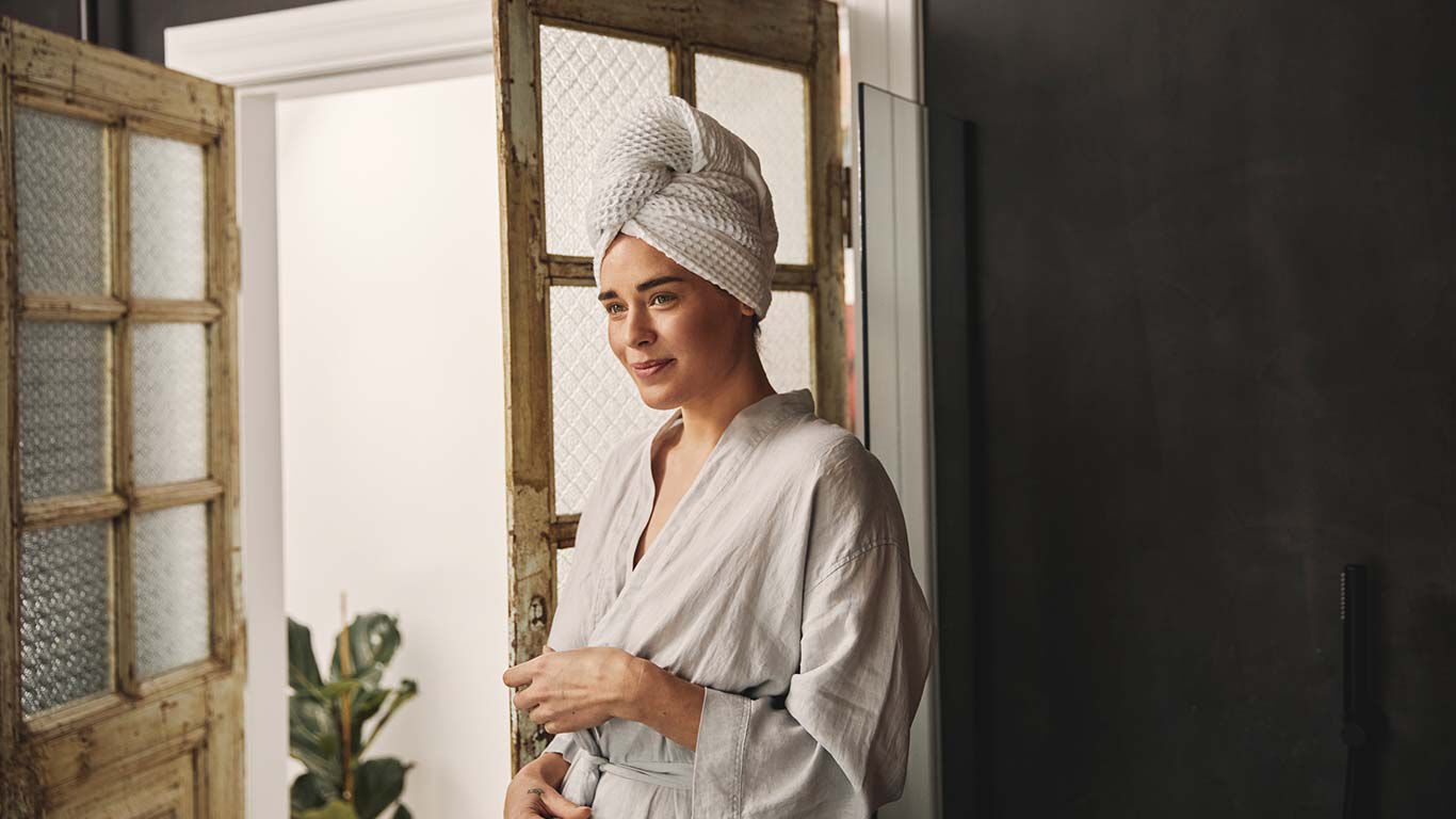 Vrouw in witte badjas en tulband staat in de badkamer.