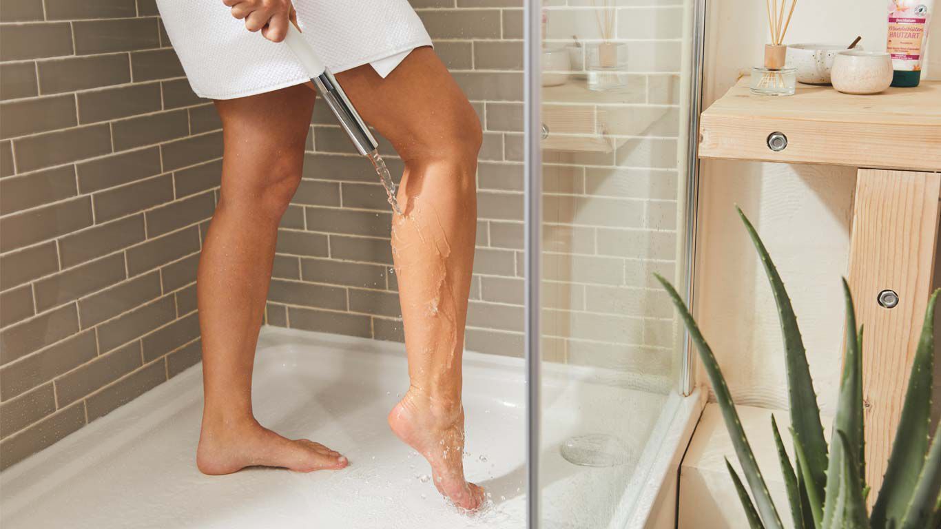 Vrouw staat onder de douche en laat water over de achterkant van haar rechterbeen stromen.