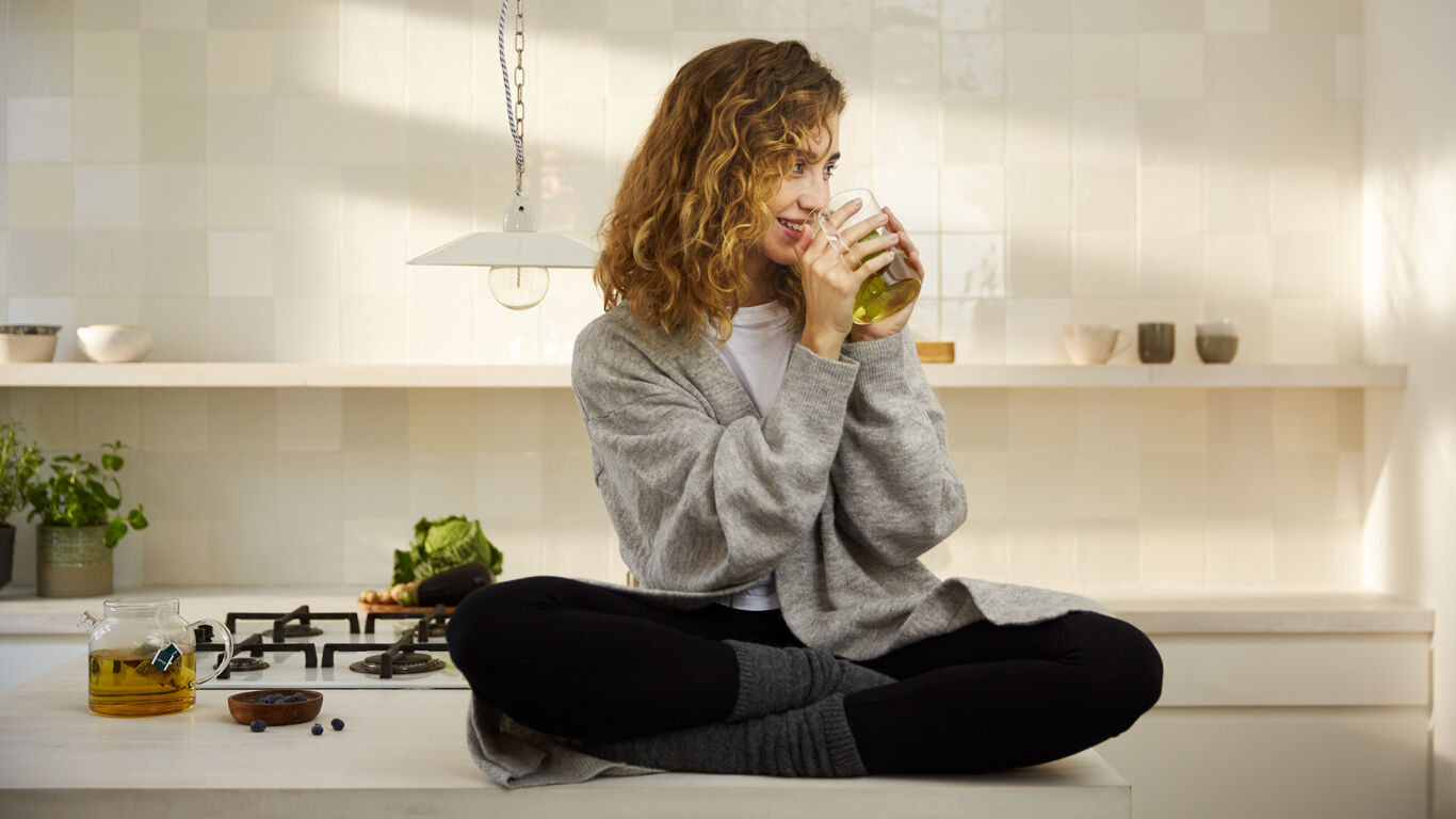 Une femme est assise en tailleur sur le comptoir de la cuisine et boit du thé dans une tasse en verre.