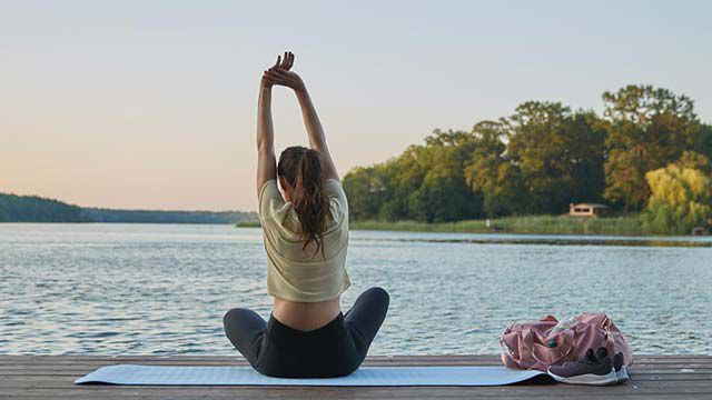 Vrouw zittend op een yogamatje aan de oever van een meer, smachtend. 