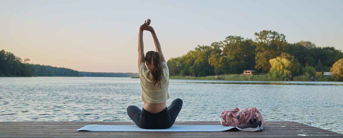 Vrouw zittend op een yogamatje aan de oever van een meer, smachtend. 