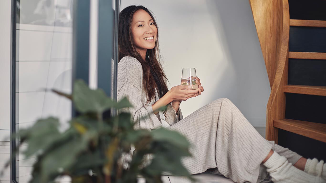 Vrouw in loungewear leunend tegen de muur met een glas water.