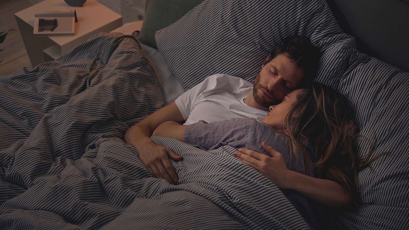 Hoeveel uur slaap hebben we nodig? Paar dat arm in arm in bed slaapt.