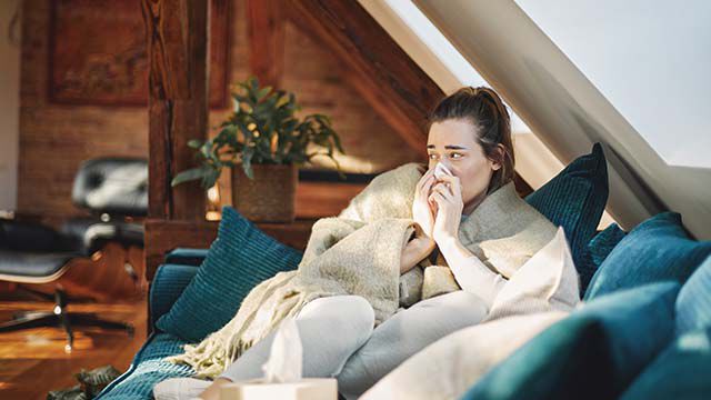 Une femme est assise sur le canapé, emmitouflée dans une couverture, et se mouche.