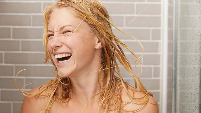 Une femme blonde sous la douche. Elle jette ses cheveux mouillés autour d'elle en riant.