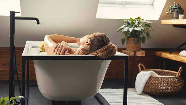 Dans une salle de bains, une femme est allongée dans une baignoire, on ne peut la voir que de dos.