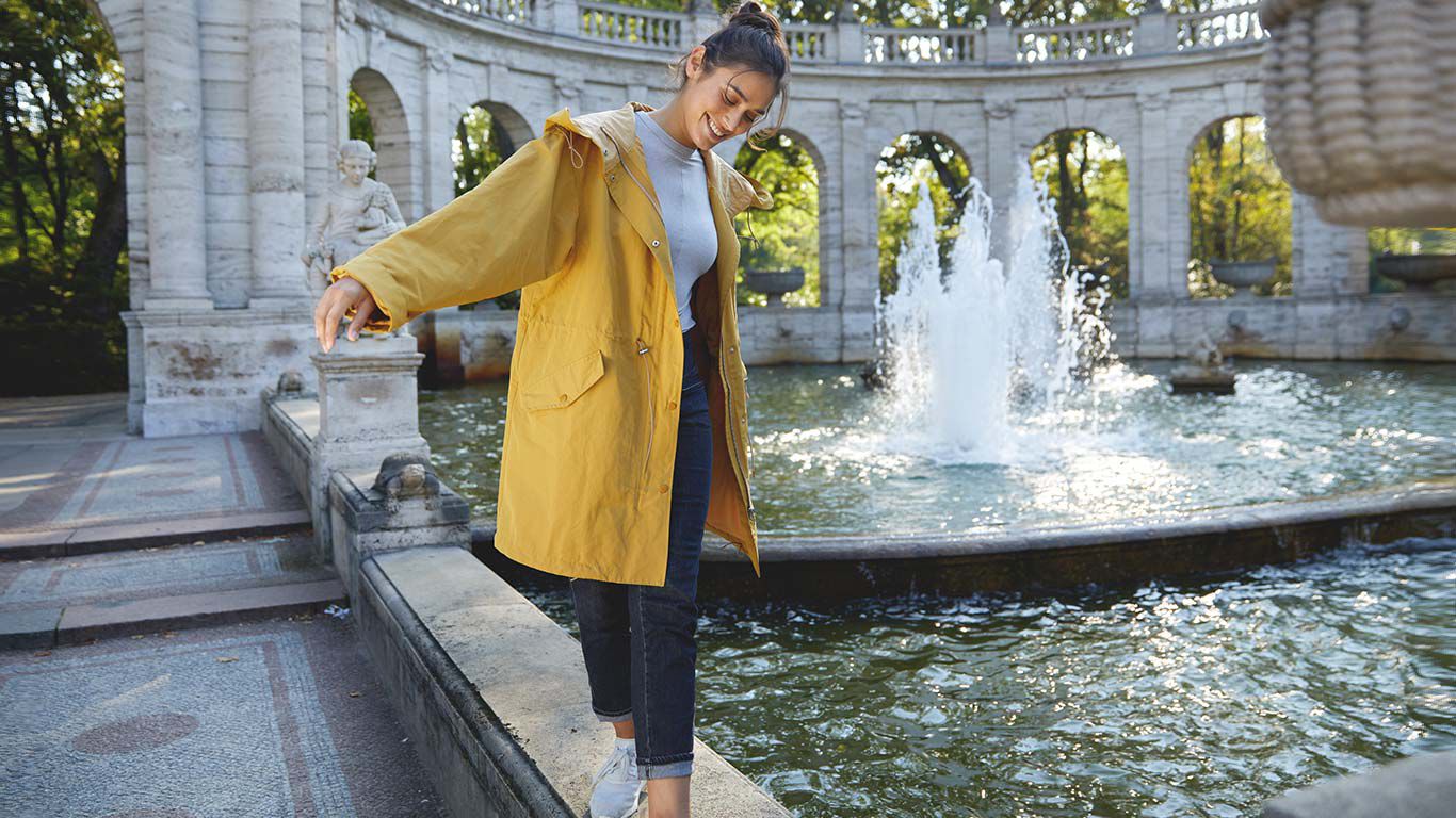Femme en imperméable jaune en équilibre sur la margelle d'une fontaine.
