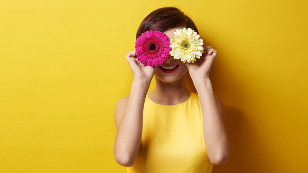 Vrouw in gele jurk tegen een gele achtergrond houdt een roze en een gele bloem voor haar ogen. 