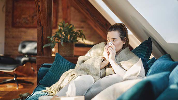 Een verkouden vrouw zit op de bank in een deken gewikkeld en snuit haar neus.