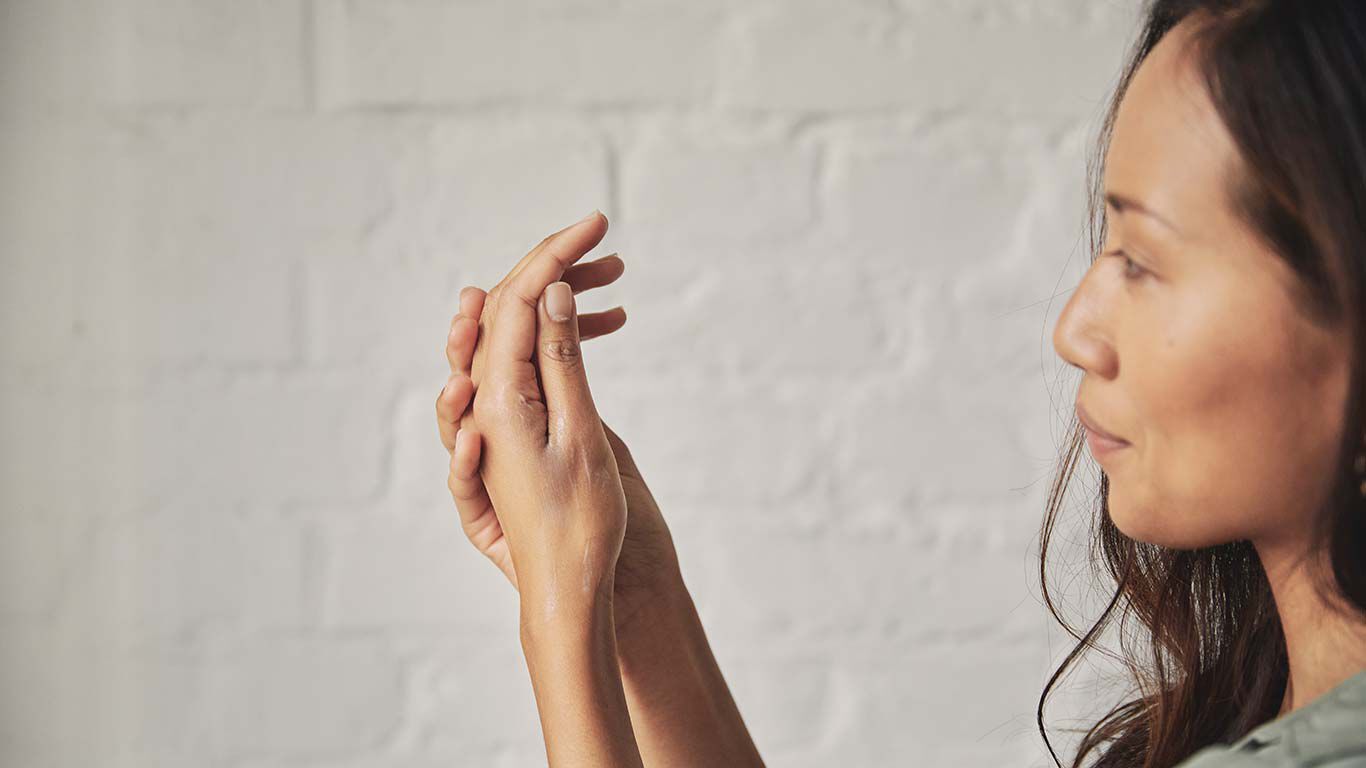 Gros plan sur une femme aux cheveux noirs et au chemisier couleur menthe en train de se passer de la crème sur les mains.