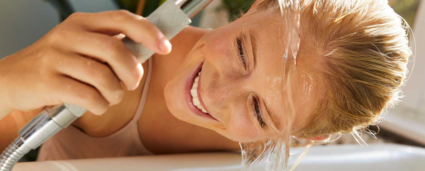 Une femme fait couler de l'eau sur son visage au-dessus de l'évier en riant.