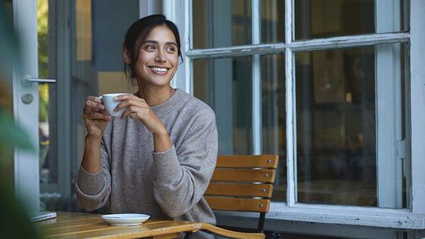 Femme assise avec une tasse de boisson chaude sur une chaise en bois, fenêtre ouverte.