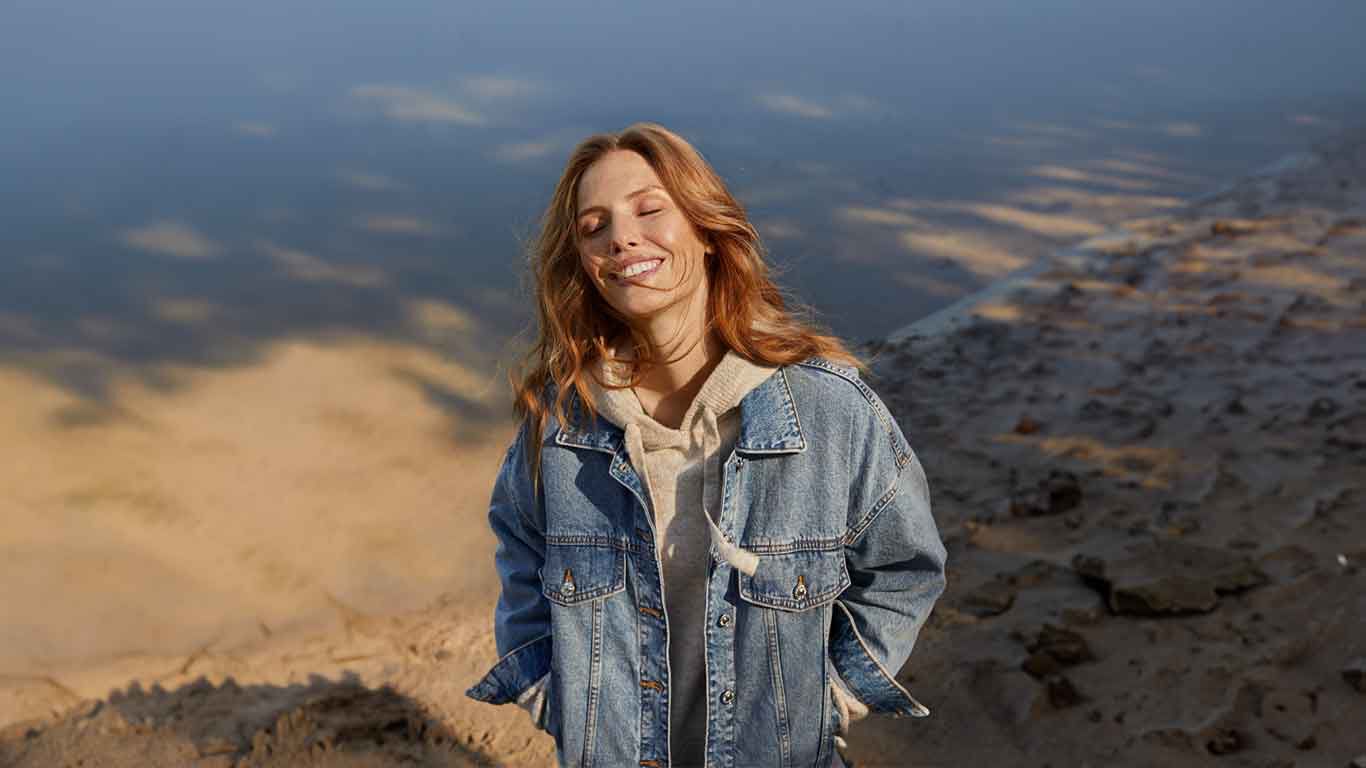 Woman in denim jacket stands laughing in nature on the beach. 