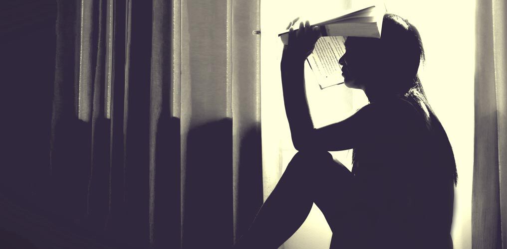 Femme assise sur le rebord d'une fenêtre, tenant un livre au-dessus de sa tête.