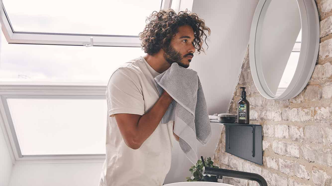 Un homme se tient dans la salle de bains devant un miroir rond et s'essuie la barbe avec une serviette. 