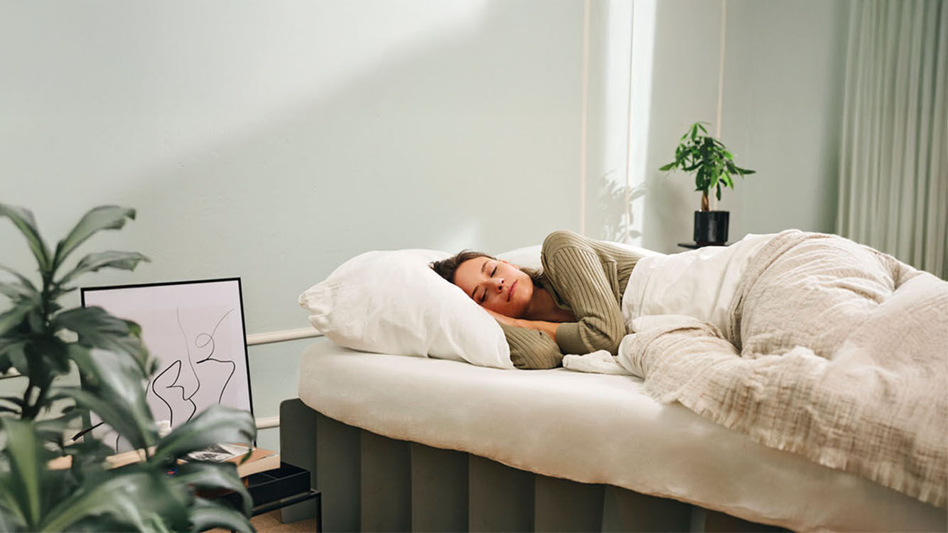 Een vrouw ligt ontspannen in bed in een licht ingerichte, natuurlijke slaapkameromgeving.