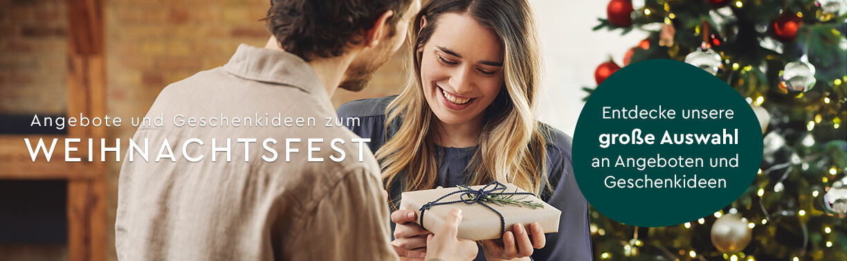 Mann übergibt Frau ein Geschenk vor dem Weihnachtsbaum