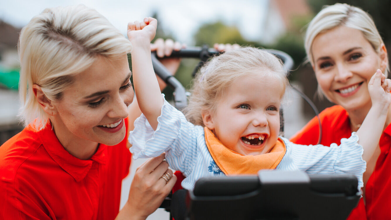 Kleines Mädchen freut sich mit hochgestreckten Armen, Zwei Frauen lachen neben ihr.