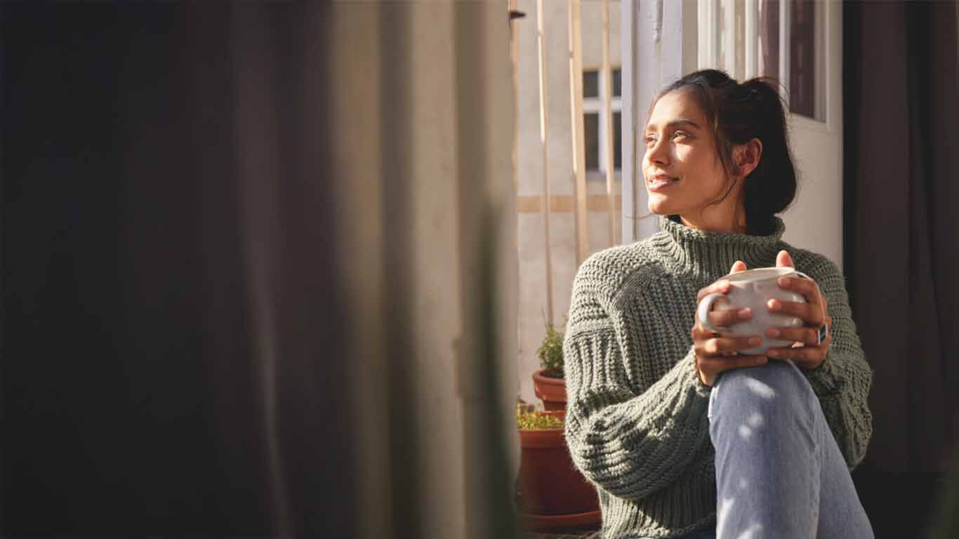 Frau sitzt auf Fensterbank mit einer Tasse Tee in der Hand und schaut nach draußen.