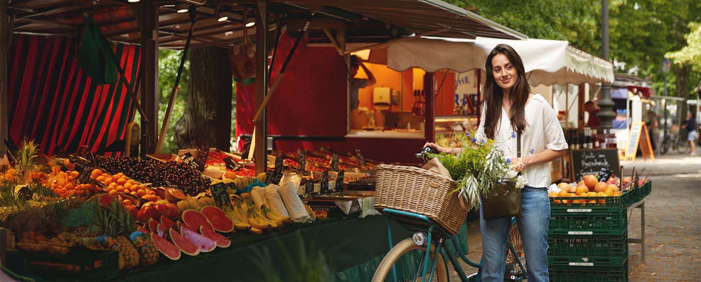 Frau mit Fahrradkorb voller Gem&uuml;se auf einem Wochenmarkt vor Obst- und Gem&uuml;sest&auml;nden.