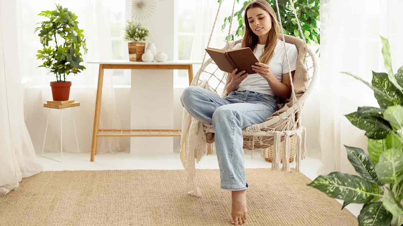 Junge Frau sitzt in einem Boho-Hängesessel in ihrer Wohnung und liest ein Buch. 