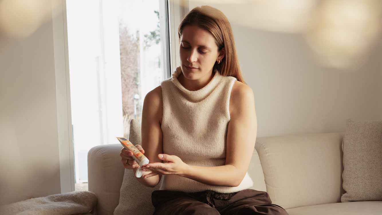 Frau sitzt auf dem Sofa und cremt ihre Hände mit Kneipp Handcreme ein. 