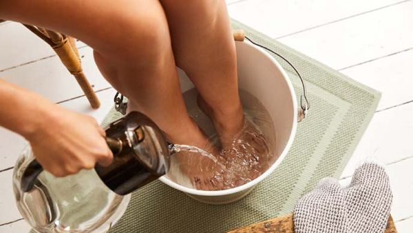 Frau taucht Füße in weisen Eimer und gießt mit einer Wasserkaraffe aus Glas Wasser nach.
