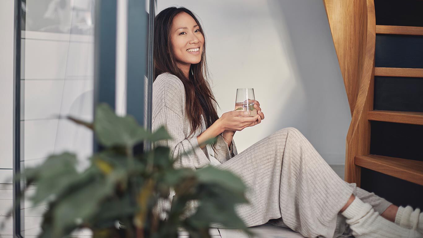 Frau in Loungewear lehnt sitzend mit einem Glas Wasser an der Wand.
