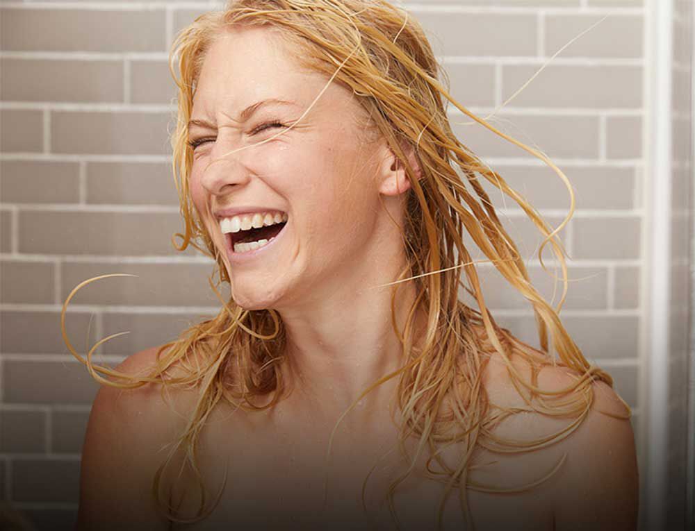 Frau mit blonden nassen Haare steht glücklich unter der Dusche. 