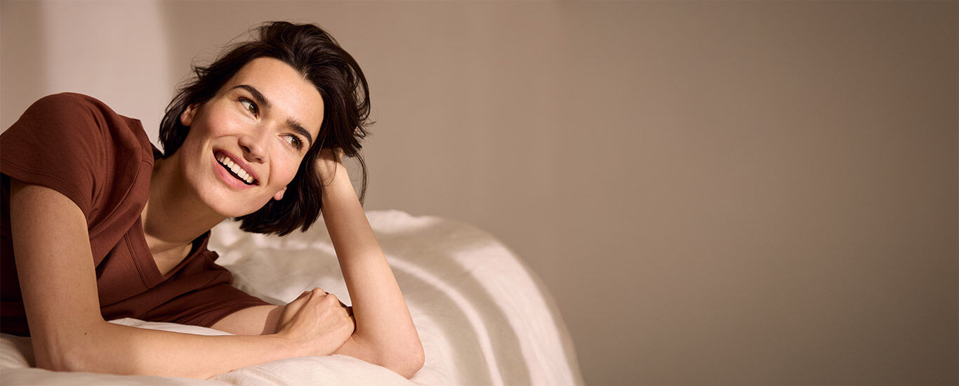 Frau liegt fr&ouml;hlich in Bauchlage im Bett und st&uuml;tzt ihren Kopf auf den H&auml;nden ab. 