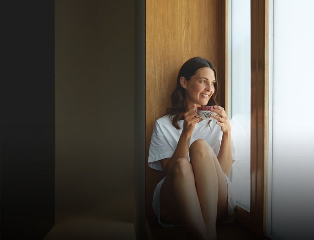 Frau sitzt mit einer Tasse am Fenster.