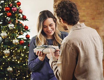 Mann übergibt Frau vor dem Weihnachtsbaum ein Geschenk.