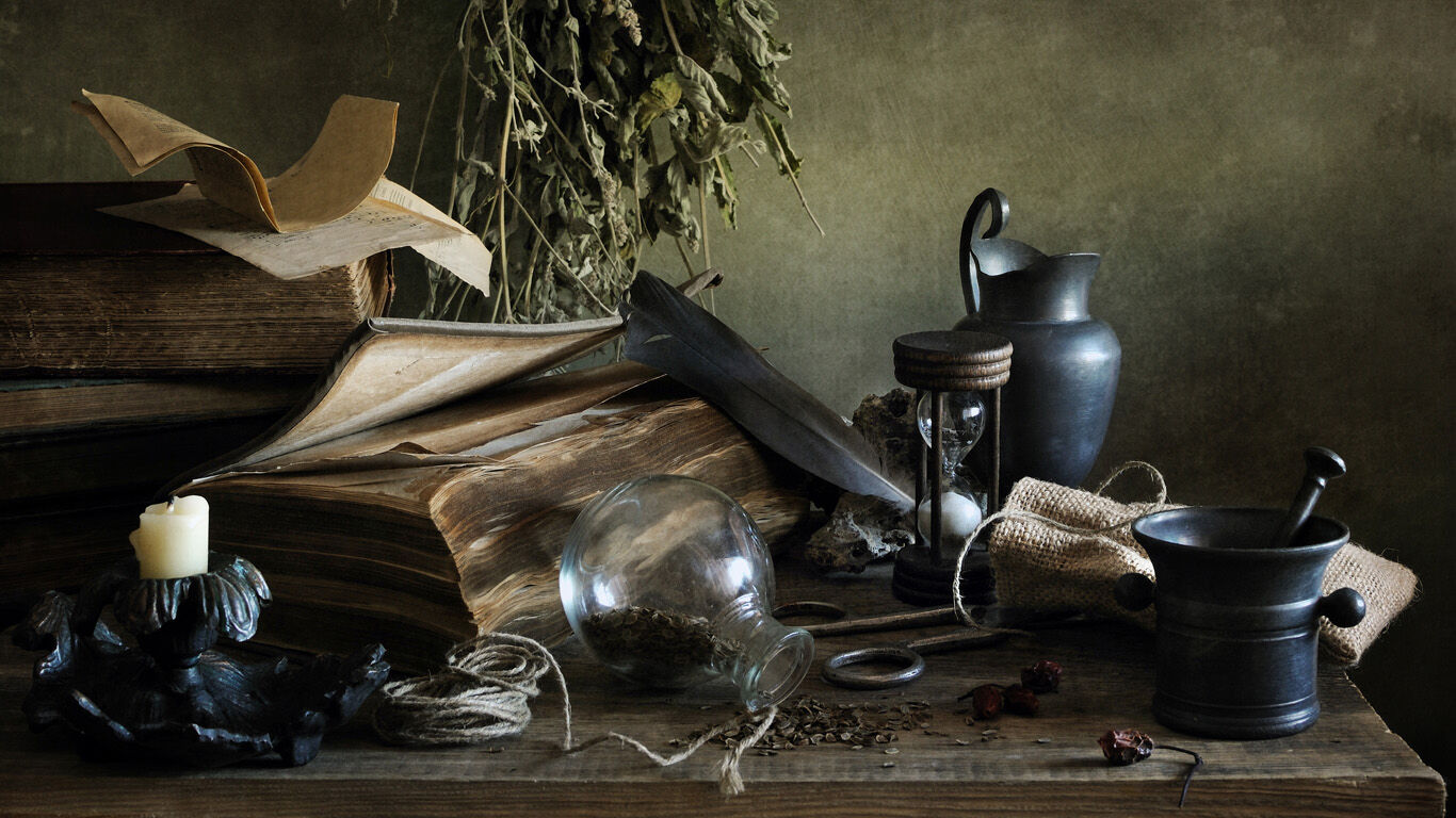 Ein alter Holztisch mit historischen Gegenständen: große Bücher, eine Sanduhr, getrocknete Kräuter, ein Mörser mit Stößel, eine Kerze und Glasflaschen