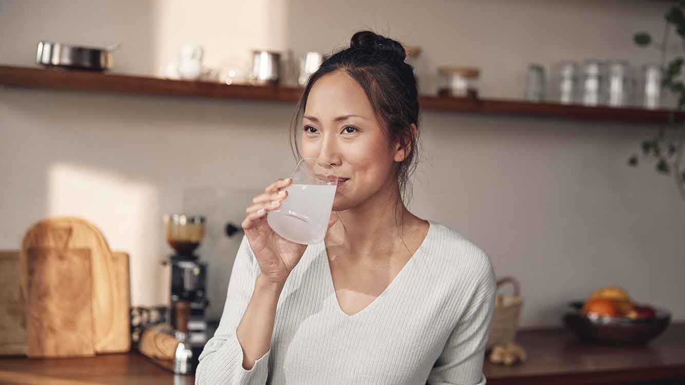  Frau steht an der Kücheninsel und trinkt ein Glas Wasser.