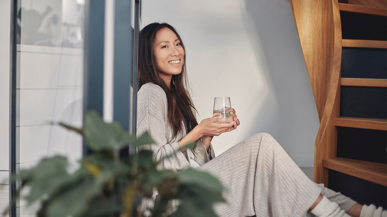 Frau sitzt angelehnt an einem Fenster mit einem Glas Wasser in der Hand.