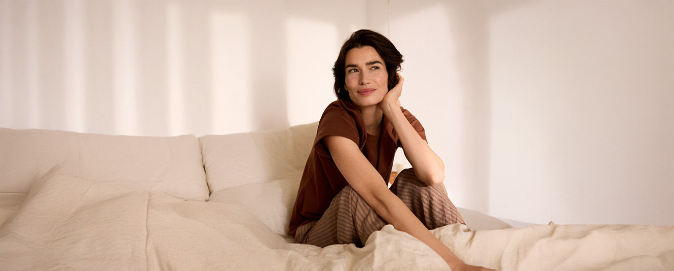 Frau sitzt entspannt in einem sonnendurchfluteten Schlafzimmer auf einem Bett mit warmen, nat&uuml;rlichen Farbt&ouml;nen.