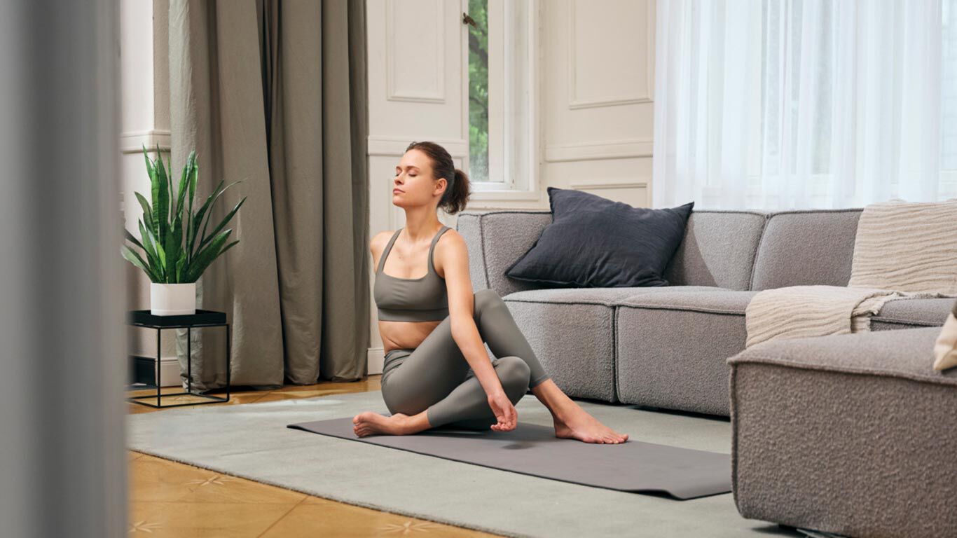 Frau f&uuml;hrt eine Yoga-Dehn&uuml;bung auf einer Yogamatte im Wohnzimmer vor einem Sofa aus.