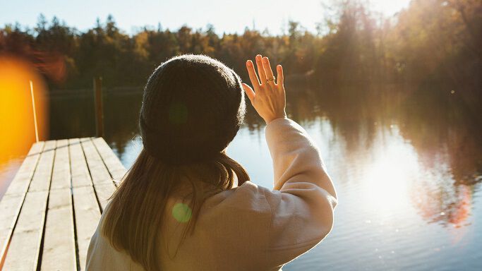 Frau steht an einem Steg und sieht der Sonne entgegen. 