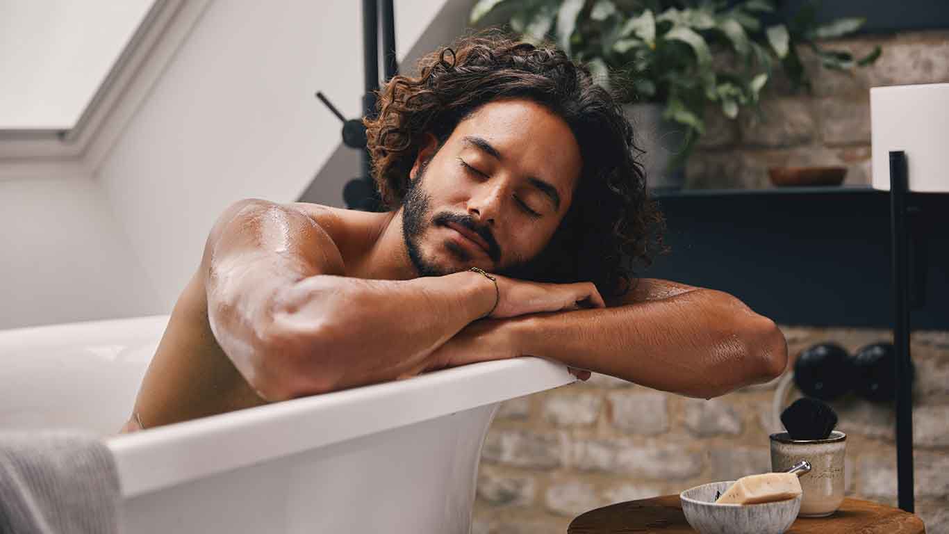 Mann entspannt in Badewanne. Er lehnt sich mit beiden Armen auf den Wannenrand und hat die Augen geschlossen.