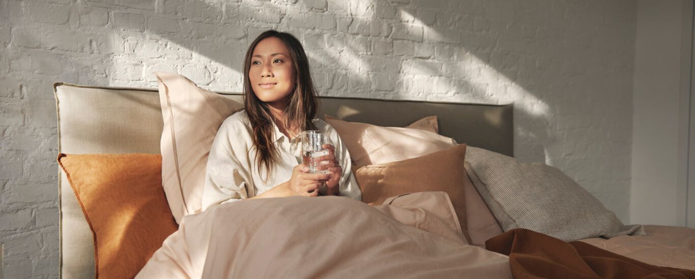  Frau sitzt mit einem Glas Wasser in den H&auml;nden aufrecht in einem Bett und die Sonne strahlt von au&szlig;en auf sie herein.