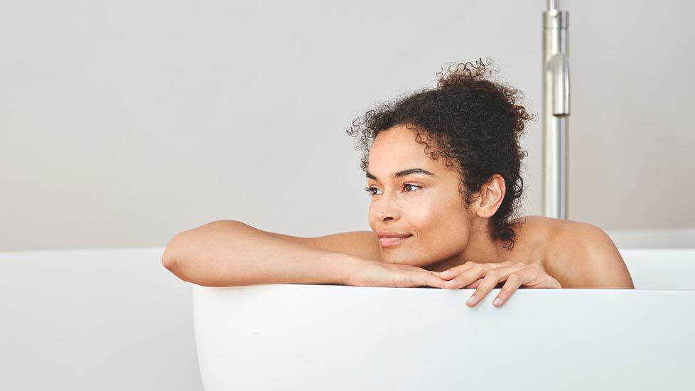 Dunkelhaarige Frau mit Locken lehnt mit Blick nach links über dem Rand einer Badewanne.