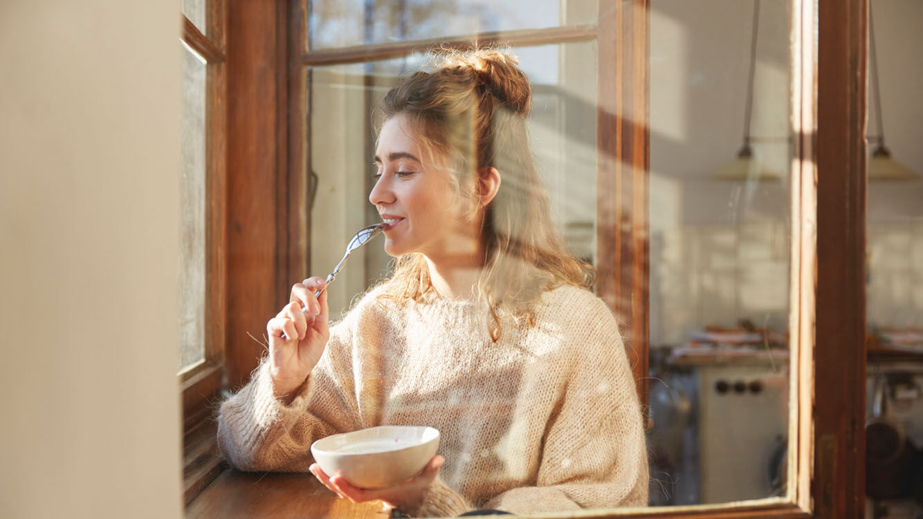 Frau sitzt mit gesunden Fr&uuml;hst&uuml;ck am Fenster mit einer Sch&uuml;ssel M&uuml;sli. Nat&uuml;rliches Tageslicht und eine entspannte Atmosph&auml;re.