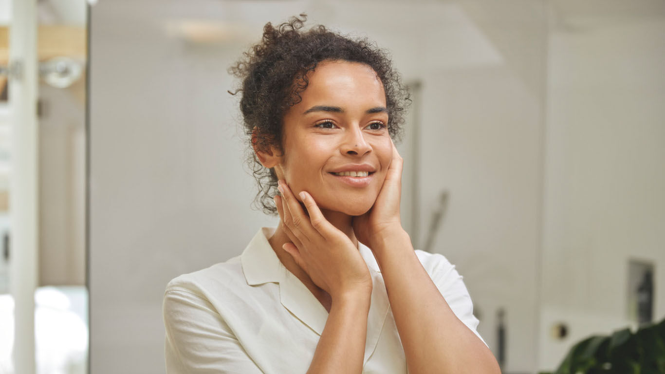 Frau steht vor einem Spiegel im steht in einem hellen Badezimmer und ber&uuml;hrt leicht ihren Hals.