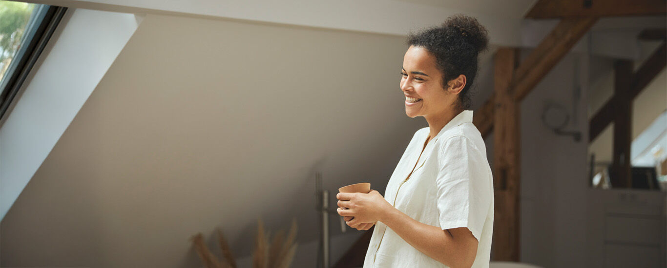 Frau steht entspannt in einem hellen, modernen Raum und h&auml;lt eine Tasse in der Hand.