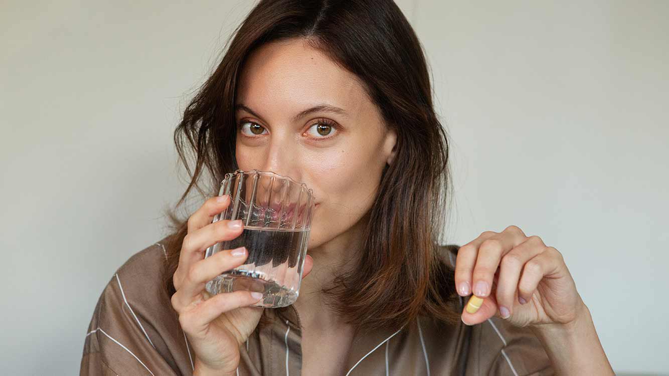 Frau trinkt aus einem Wasserglas. In der Hand hält sie ein Nahrungsergänzungsmittel. 