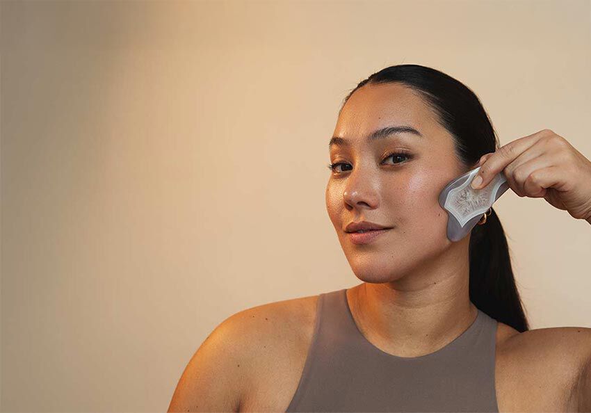 Schwarzhaarige Frau im Top führt einen Gua Sha Stein über ihr Gesicht. 