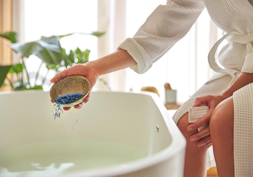 Frau im Bademantel kippt Badesalz in Badewanne.