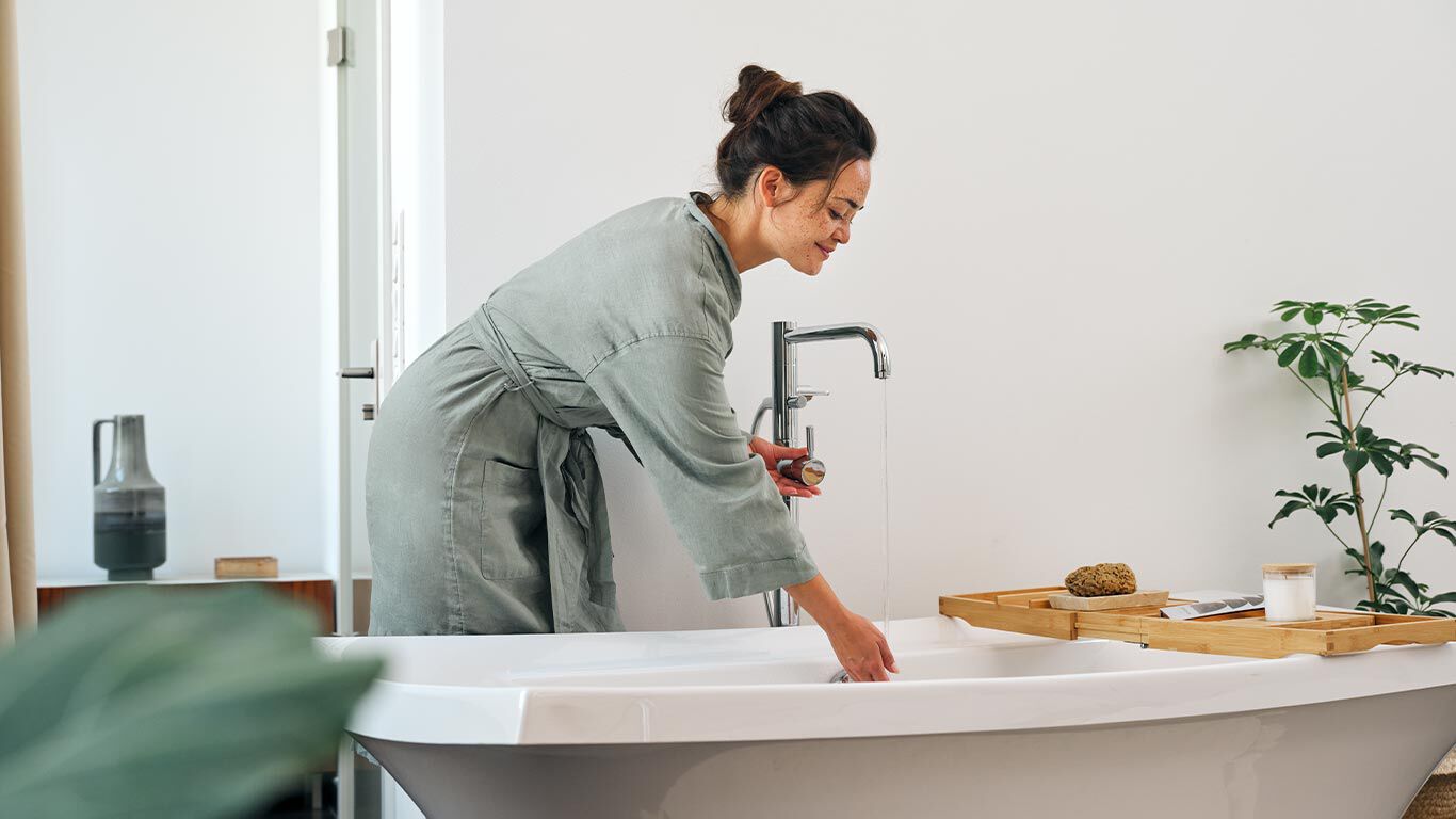 Frau in Bademantel testet Wassertemperatur in Badewanne. 